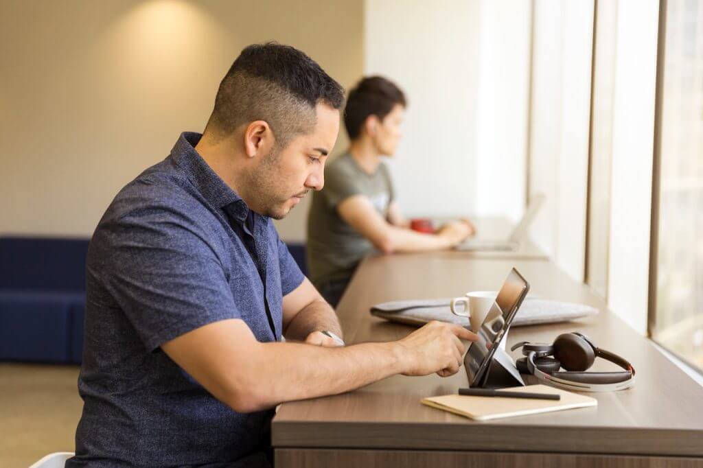 Hombre trabajando en una tablet en un espacio de coworking, con otro trabajador de fondo usando una laptop.